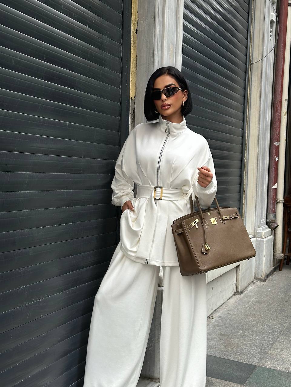 Woman in a white outfit with a brown handbag standing against a gray wall.